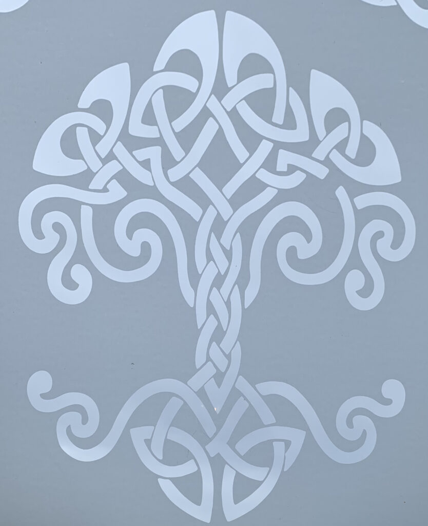 A celtic knot design that resembles a giant tree. The knots on top create the foliage and expansive branches of a large tree. There is a braided trunk, and a visible root system that has three definable sections. 