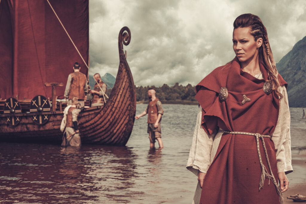 This image shows a viking woman wearing clothes that are an accurate portrayal of what the vikings would wear. The woman has a simple fiber shift, with a wool cloak over it. It is tied with a rope belt and she has brooches. 